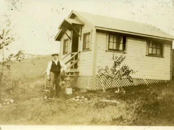 Sepia photograph of a man standing beside one of the camp's first wooden cottages