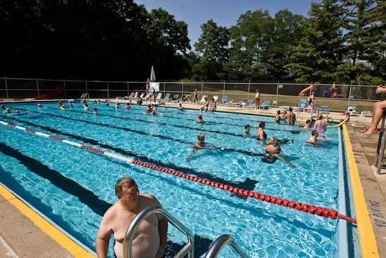 Swimmers spread across the outdoor lap pool at Illinois Turners Camp on a bright afternoon, with lane ropes in the water and deck chairs along the poolside