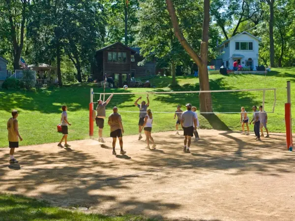 Members playing a volleyball game on the sand court with cottages on the hillside behind