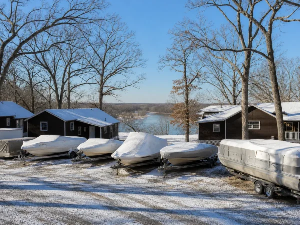 Boats under white winter covers on their trailers among bare oaks, with brown-sided cottages behind and the Fox River below the bluff, at Illinois Turner Camp in winter
